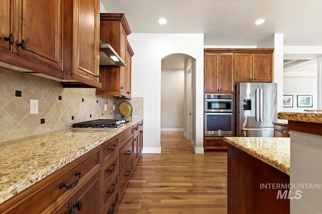 kitchen featuring stainless steel appliances, arched walkways, wall chimney exhaust hood, light stone counters, and baseboards