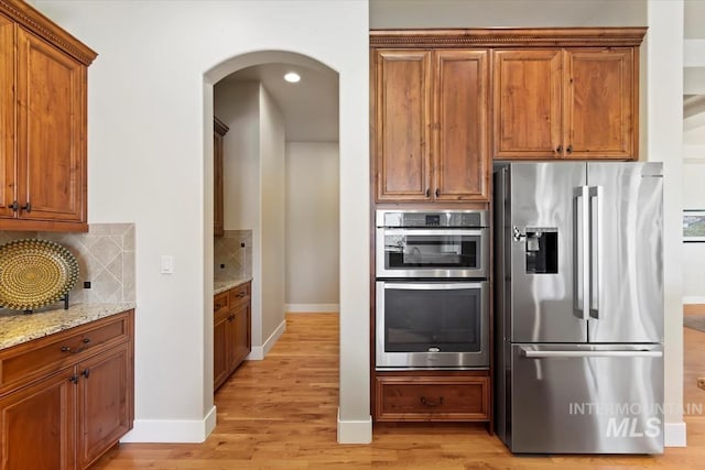 kitchen with stainless steel appliances, light stone counters, light wood finished floors, backsplash, and arched walkways