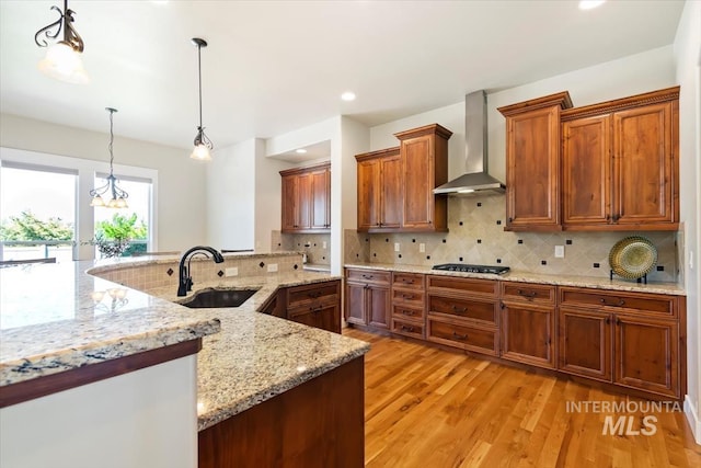 kitchen featuring wall chimney range hood, a sink, gas stovetop, brown cabinetry, and light wood-style floors