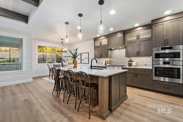 kitchen with dark brown cabinets, hanging light fixtures, wall chimney exhaust hood, and an island with sink