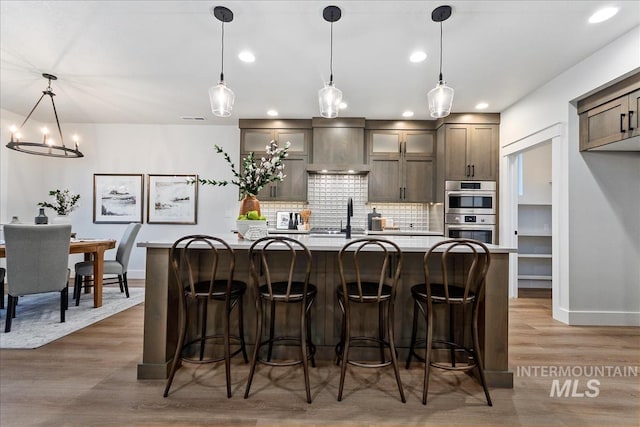 kitchen featuring dark wood-type flooring, wall chimney range hood, an island with sink, and hanging light fixtures