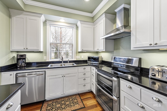 kitchen featuring white cabinets, dark countertops, stainless steel appliances, wall chimney range hood, and a sink