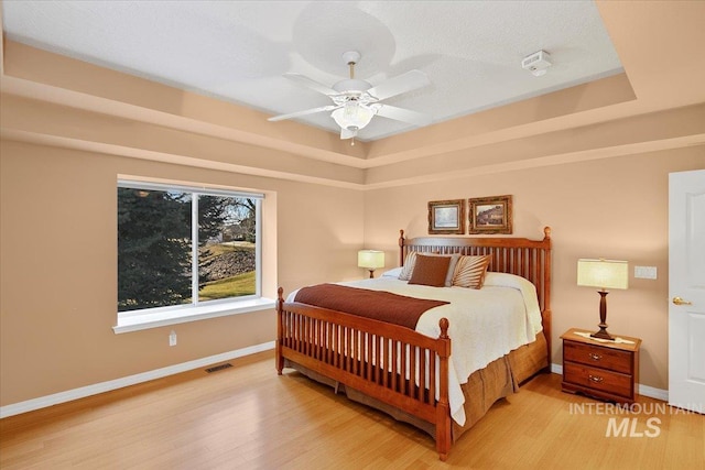 bedroom with visible vents, baseboards, ceiling fan, light wood-style flooring, and a tray ceiling