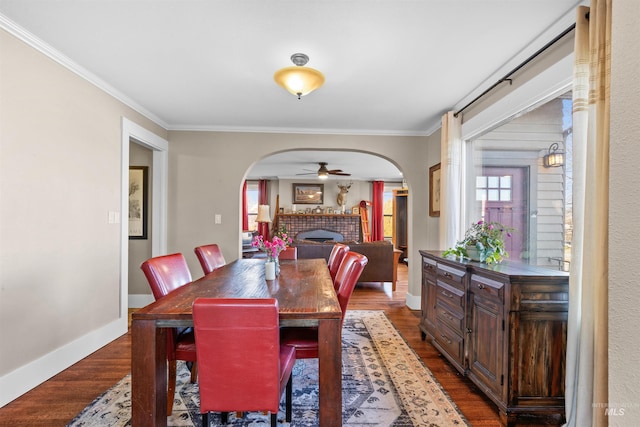 dining area featuring arched walkways, dark wood finished floors, a tiled fireplace, ornamental molding, and baseboards