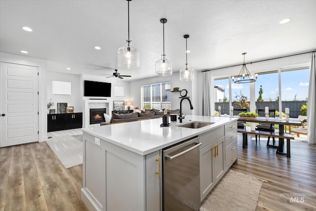 kitchen with light wood finished floors, a kitchen island with sink, a warm lit fireplace, a sink, and stainless steel dishwasher