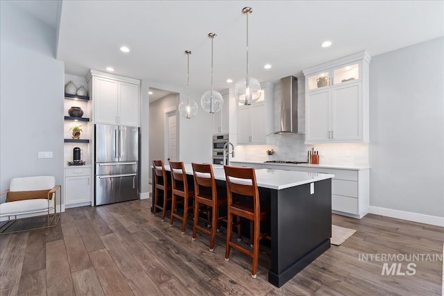 kitchen featuring dark wood-style flooring, light countertops, appliances with stainless steel finishes, wall chimney exhaust hood, and an island with sink