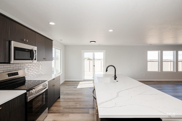 kitchen with light wood-style flooring, a kitchen island with sink, a sink, tasteful backsplash, and appliances with stainless steel finishes