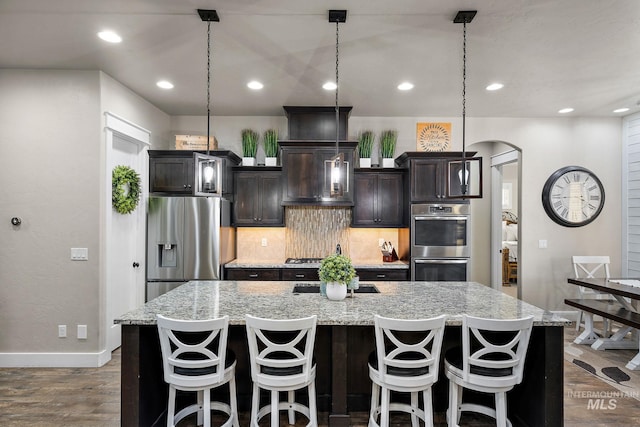 kitchen with a center island with sink, arched walkways, decorative backsplash, dark wood finished floors, and appliances with stainless steel finishes