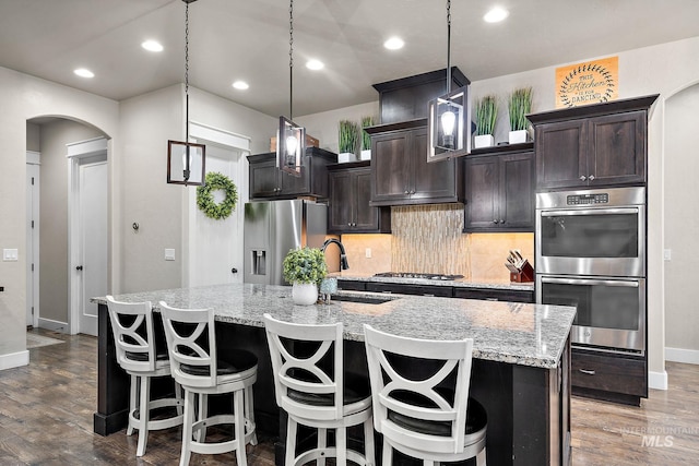 kitchen with arched walkways, an island with sink, appliances with stainless steel finishes, wood finished floors, and a sink