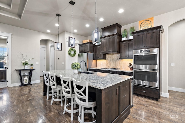 kitchen featuring arched walkways, dark wood finished floors, an island with sink, stainless steel appliances, and a sink