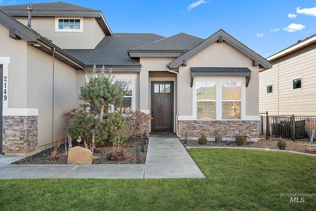 doorway to property featuring roof with shingles, stucco siding, a lawn, fence, and stone siding