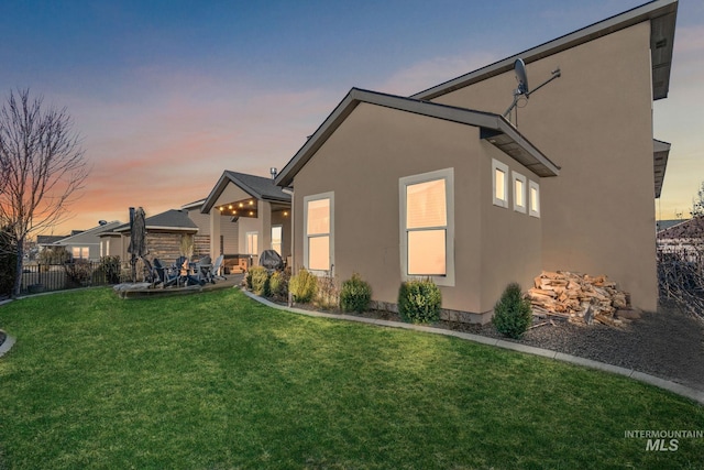 back of house at dusk with stucco siding, a patio area, fence, and a yard