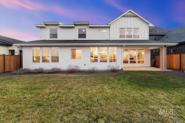 back of house at dusk featuring board and batten siding, fence, a patio, and a lawn