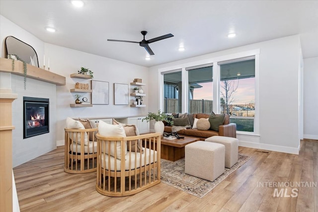 living room with ceiling fan, light wood finished floors, a tiled fireplace, and baseboards