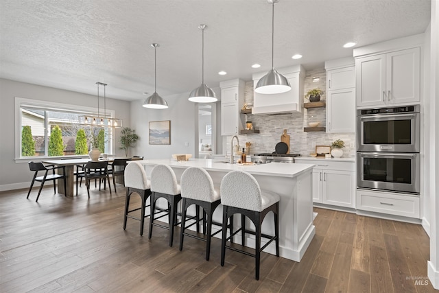 kitchen with dark wood-style floors, a center island with sink, tasteful backsplash, light countertops, and double oven