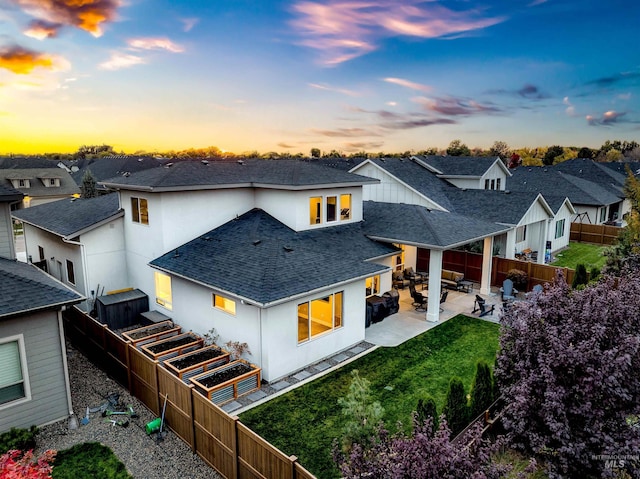 back of house at dusk featuring roof with shingles, a yard, a patio, a residential view, and a fenced backyard