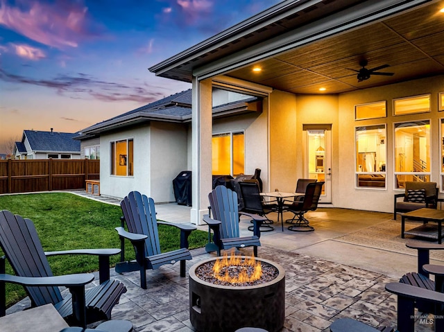 patio terrace at dusk with a fire pit, a yard, ceiling fan, and fence