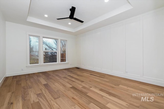 empty room featuring a decorative wall, light wood finished floors, ceiling fan, recessed lighting, and a tray ceiling