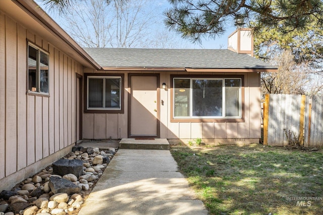 view of exterior entry with fence, a yard, a shingled roof, a chimney, and board and batten siding