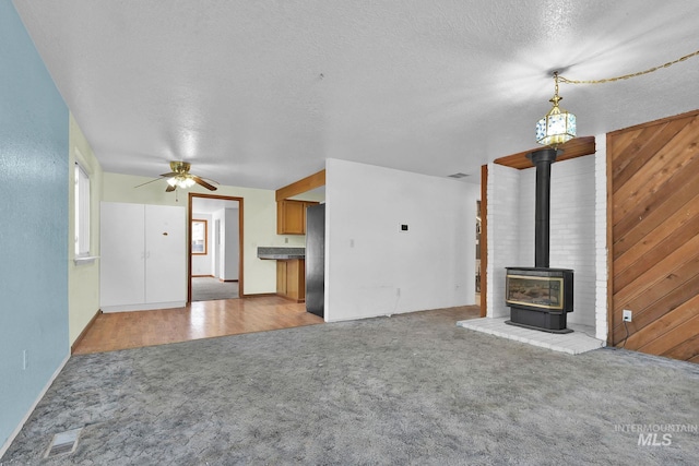 unfurnished living room with a wood stove, ceiling fan, a textured ceiling, light colored carpet, and wood walls