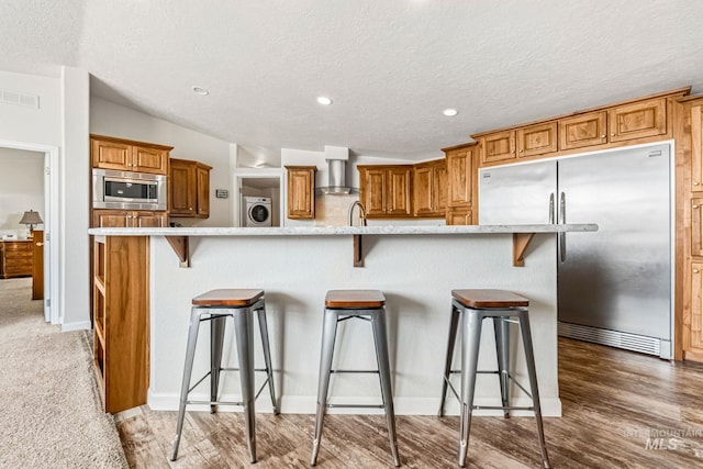 4459 E Deer Flt, Kuna, ID 83634 - kitchen with appliances with stainless steel finishes, a breakfast bar area, brown cabinetry, a textured ceiling, and wall chimney exhaust hood