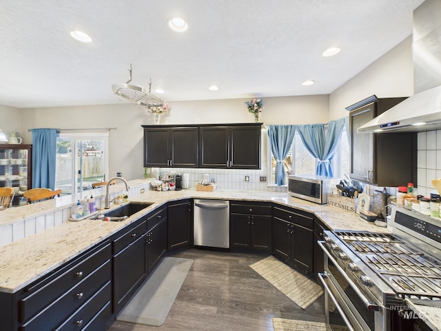 kitchen with a sink, backsplash, stainless steel appliances, a peninsula, and wall chimney range hood