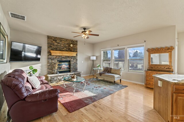 living room featuring light wood-style flooring, ceiling fan, baseboards, a textured ceiling, and a stone fireplace