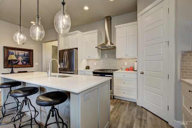 kitchen with sink, wall chimney range hood, an island with sink, white cabinets, and appliances with stainless steel finishes