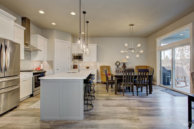 kitchen featuring wall chimney exhaust hood, hanging light fixtures, stainless steel appliances, light hardwood / wood-style flooring, and an island with sink