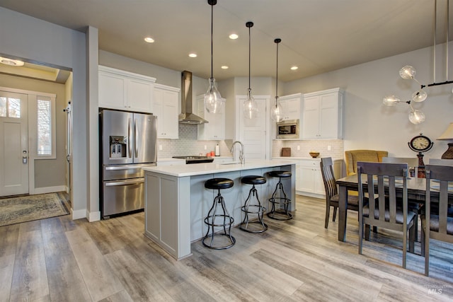 kitchen featuring appliances with stainless steel finishes, a kitchen island with sink, wall chimney range hood, white cabinets, and light hardwood / wood-style floors