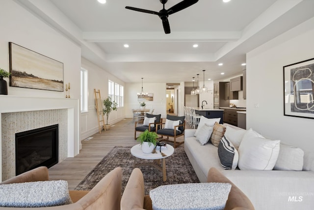 living room with recessed lighting, beamed ceiling, light wood-type flooring, a glass covered fireplace, and ceiling fan with notable chandelier
