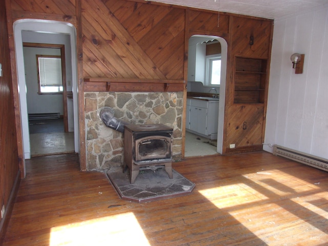 unfurnished living room featuring arched walkways, wood-type flooring, baseboard heating, a wood stove, and wood walls