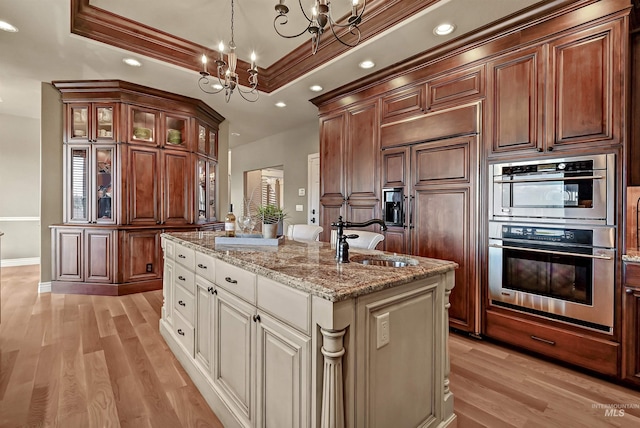 kitchen with double oven, a raised ceiling, a kitchen island with sink, and a sink