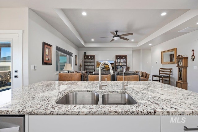 kitchen with light stone counters, a tray ceiling, sink, and a center island with sink