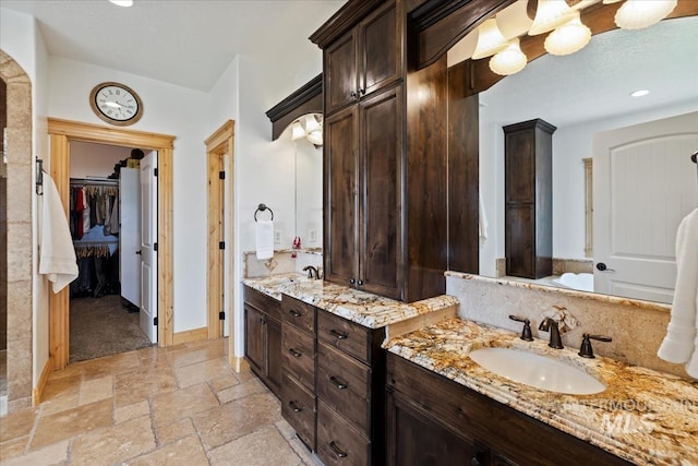 full bathroom featuring stone tile flooring, baseboards, two vanities, a spacious closet, and a sink