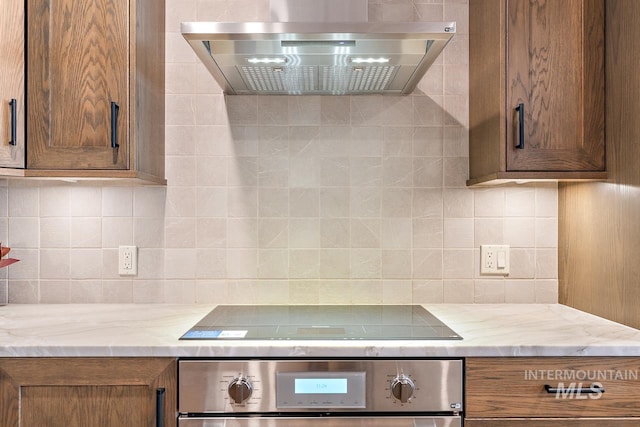 kitchen with tasteful backsplash, black electric stovetop, light stone counters, wall oven, and wall chimney range hood