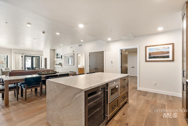 kitchen with a kitchen island, stainless steel microwave, beverage cooler, light stone countertops, and light wood-type flooring