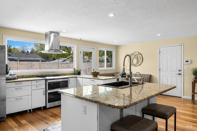 kitchen featuring a breakfast bar, a sink, a textured ceiling, white cabinetry, and island range hood