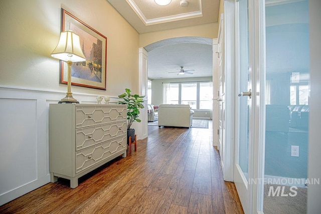 hall featuring arched walkways, wainscoting, a decorative wall, and wood finished floors