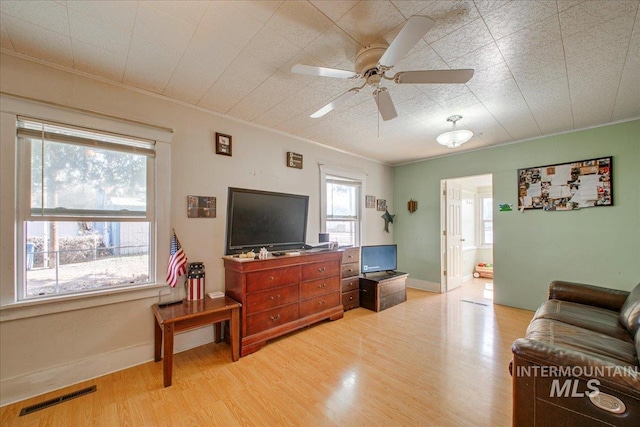 living area with visible vents, baseboards, ceiling fan, light wood-type flooring, and ornamental molding