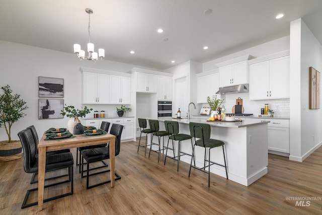 kitchen with tasteful backsplash, white cabinetry, hanging light fixtures, a kitchen island with sink, and light wood-type flooring