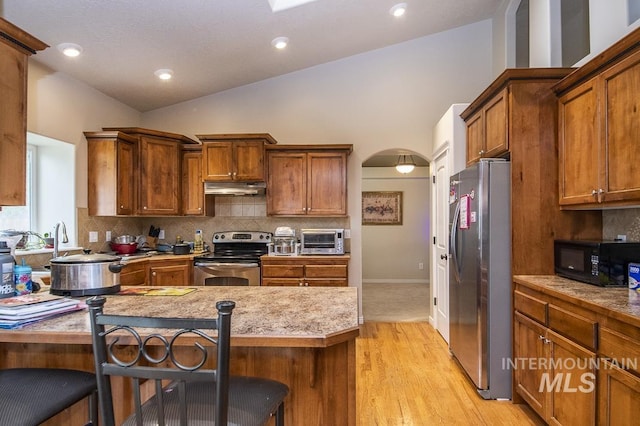 kitchen with arched walkways, decorative backsplash, appliances with stainless steel finishes, under cabinet range hood, and a kitchen breakfast bar