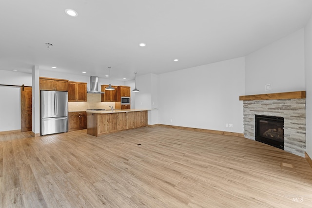 kitchen with a barn door, wall chimney exhaust hood, light hardwood / wood-style floors, stainless steel appliances, and hanging light fixtures