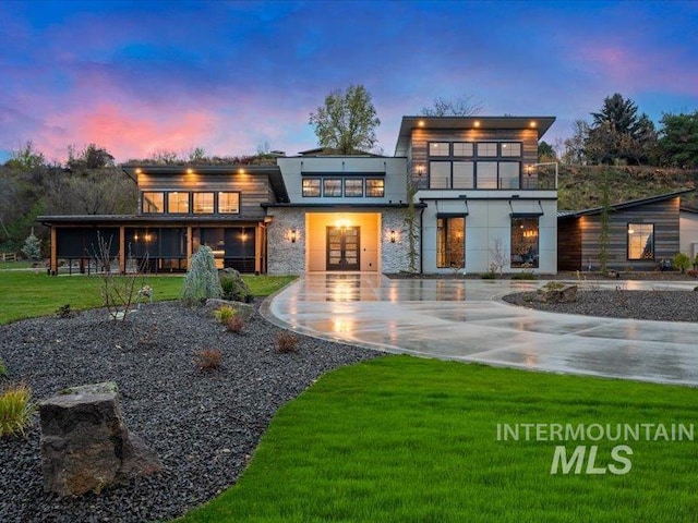 back of house at dusk with french doors, stone siding, curved driveway, and a yard