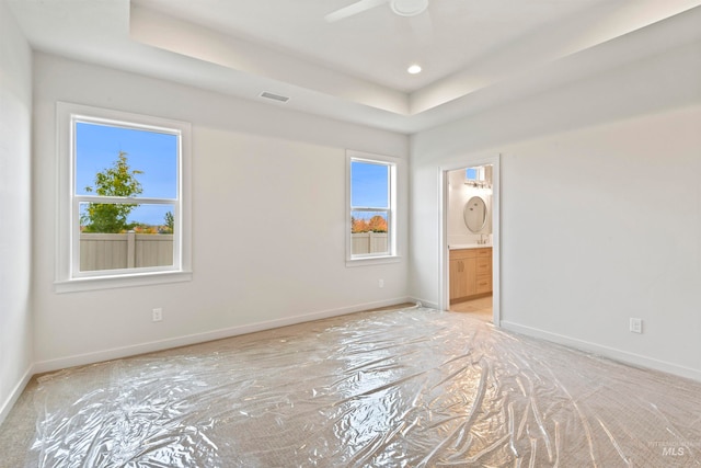 unfurnished bedroom with multiple windows, ensuite bathroom, a tray ceiling, and ceiling fan