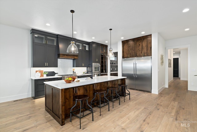 kitchen featuring hanging light fixtures, appliances with stainless steel finishes, light wood-type flooring, and a kitchen island with sink