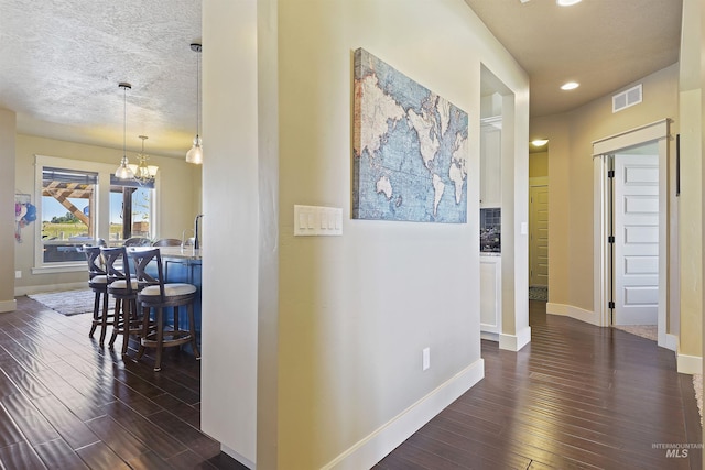 corridor with dark wood finished floors, visible vents, an inviting chandelier, a textured ceiling, and baseboards