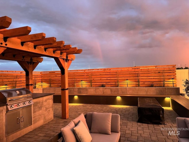 patio terrace at dusk with area for grilling, an outdoor kitchen, a fenced backyard, and a pergola
