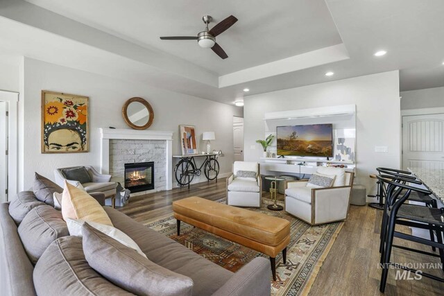 living area featuring ceiling fan, a fireplace, wood finished floors, recessed lighting, and a tray ceiling