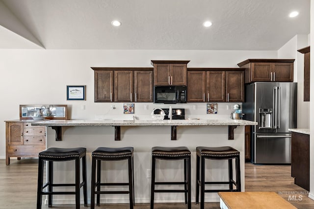 kitchen featuring black microwave, high quality fridge, dark brown cabinetry, a breakfast bar, and wood finished floors